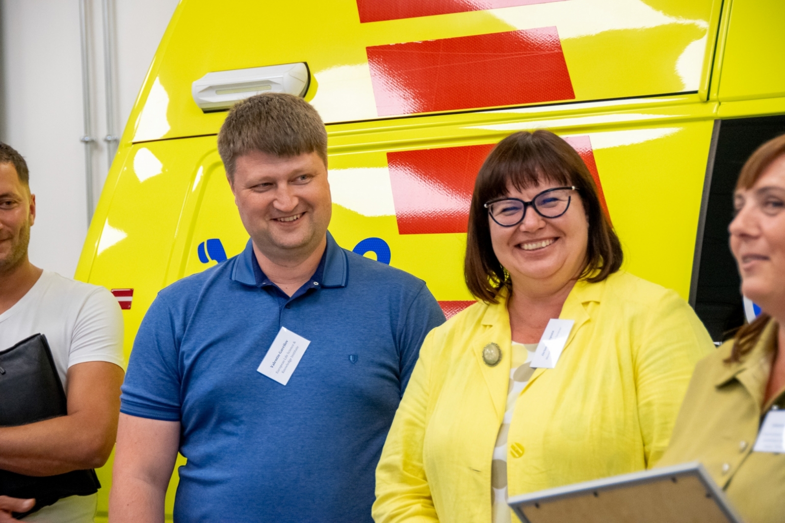 vīrietis zilā kreklā un sieviete dzeltenā žaketē smaida. Fonā dzeltens operatīvais auto